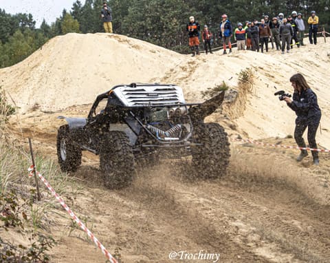 Czarny buggy wjeżdża na piaszczystą wydmę, obok operatorka kamery, na grzbiecie kibice