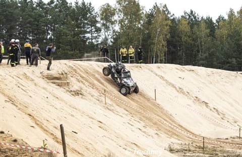 Buggy zjeżdża stromym piaszczystym stokiem, na górze obserwują strażacy i kibice