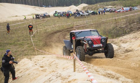 Czerwono-czarny buggy pokonuje piaszczysty zakręt przy taśmach, obok fotograf z aparatem