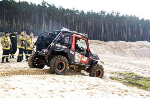 Czarny buggy z czerwonymi drzwiami na piachu, obok grupa strażaków w mundurach
