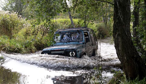 Ciemnoturkusowa terenówka z listwą LED rozbija spienioną falę w leśnym strumieniu