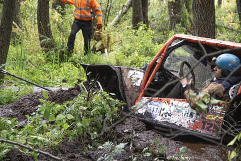 Kierowca w niebieskim kasku prowadzi pomarańczowy buggy przez błoto i zarośla