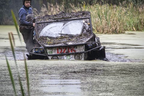 Zawodnik w woderach prowadzi zabłoconą terenówkę z napisem DZIK przez staw