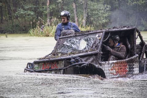 Oblepiony błotem buggy brnie przez głęboką wodę, członek załogi w kasku stoi przy kabinie