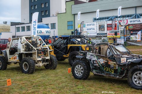 Biały buggy i czarny pojazd z klatką w parku maszyn przed halą