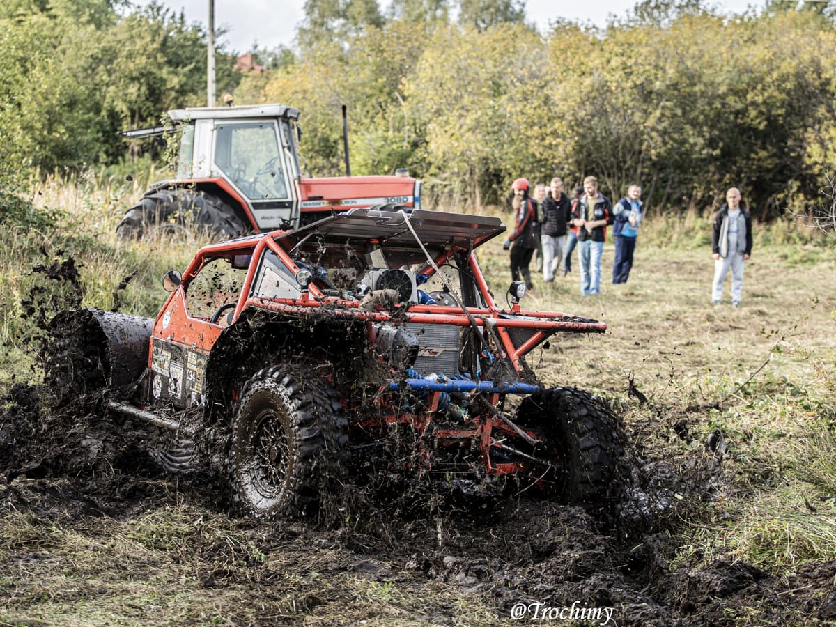 Czerwony buggy z klatką rozbryzguje błoto, w tle widzowie i ciągnik