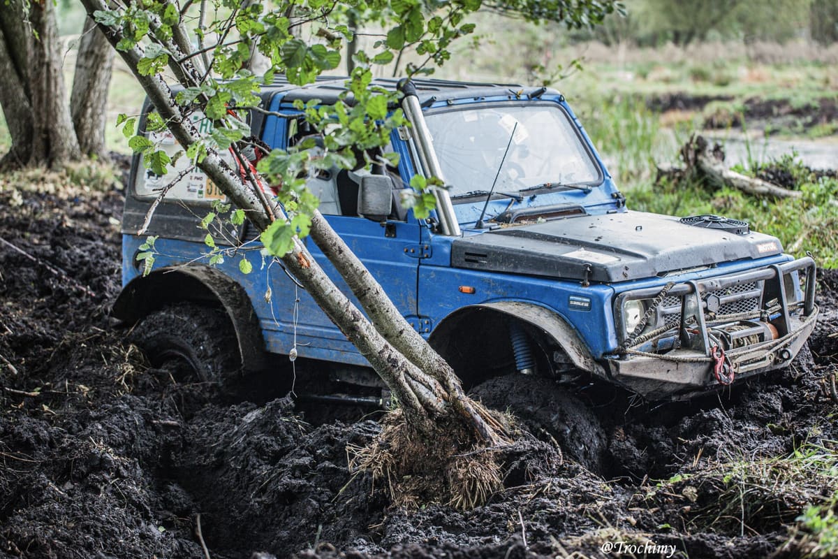 Niebieskie Suzuki Samurai wjeżdża w błoto, przewracając młode drzewo przed maską