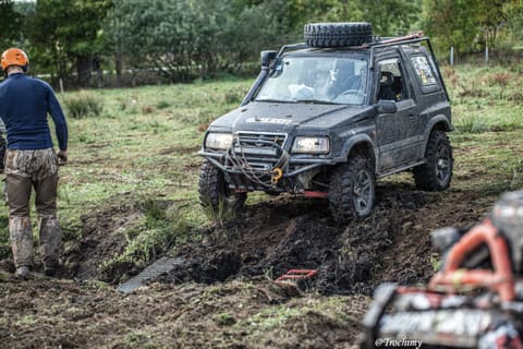 Grafitowe Suzuki Vitara wspina się z rowu, obok idzie zawodnik w pomarańczowym kasku