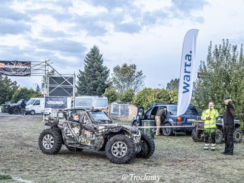 Kamuflażowy buggy na trawiastym placu bazy przy flagach sponsora i obsłudze