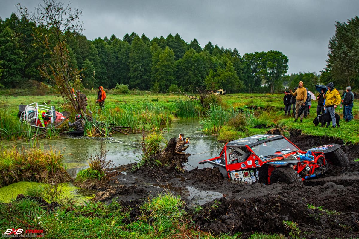 Czerwony buggy utknął w błocie nad zarośniętym stawem, widzowie obserwują z łąki