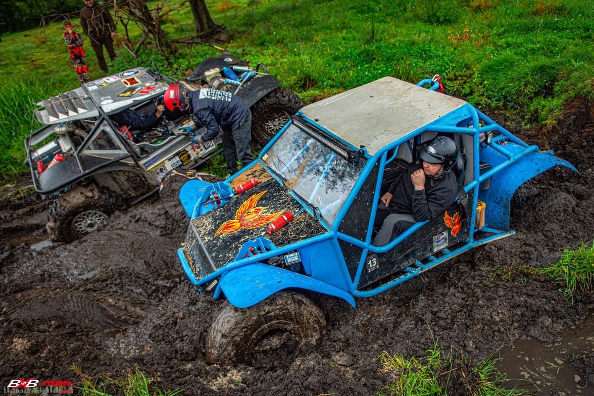 Niebieski buggy z klatką grzęźnie w błocie, za nim załoga pracuje przy srebrnym pojeździe
