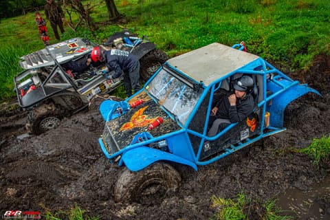 Niebieski buggy z klatką grzęźnie w błocie, za nim załoga pracuje przy srebrnym pojeździe