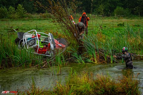 Srebrny buggy przewrócony w zarośniętym stawie, zawodnik brodzi w wodzie z liną