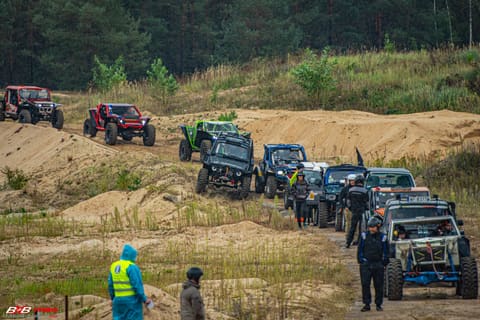 Kolumna terenówek czeka w kolejce na piaszczystej trasie, obok osoba w odblaskowej kamizelce