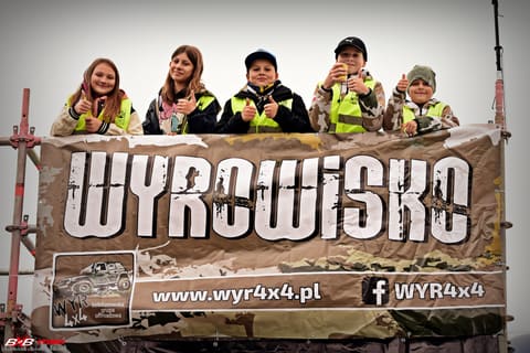 Pięcioro dzieci w odblaskowych kamizelkach pokazuje kciuki nad banerem WYRowisko na rusztowaniu