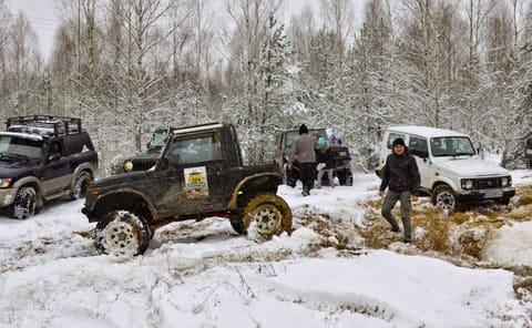 Zabłocony Suzuki Samurai z numerem 206 na zaśnieżonej przeprawie, obok uczestnicy i terenówki